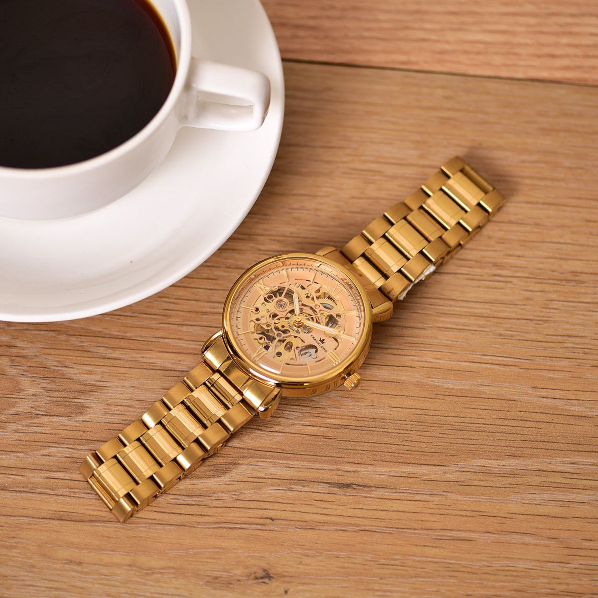 Franklord Mechanis Regal Gold Watch elegantly placed on a wooden surface beside a cup of coffee, showcasing its premium skeleton dial and gold bracelet.